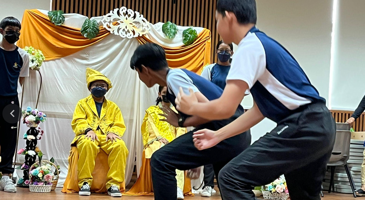 Two boys perform in front of a child seated in yellow costume on a decorated stage with orange and white drapes.